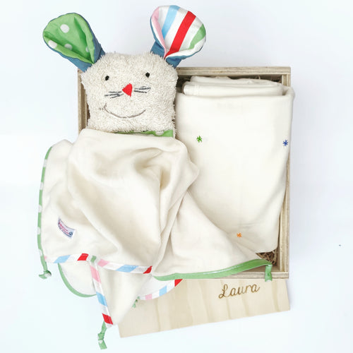 Hier siehst du ein beiges Stofftier mit bunten Ohren und einen Körper aus einem kleinem Tuch in einer Holzbox liegend. Neben dem Stofftier liegt eine zusammengefaltetete Decke mit bunten Sternchen eingehnäht. 