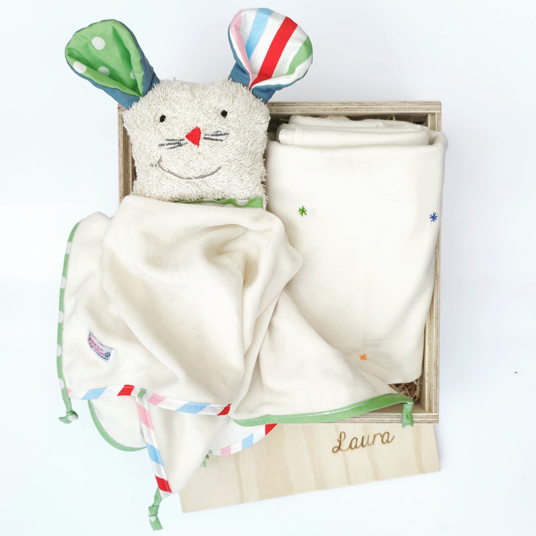 Hier siehst du ein beiges Stofftier mit bunten Ohren und einen Körper aus einem kleinem Tuch in einer Holzbox liegend. Neben dem Stofftier liegt eine zusammengefaltetete Decke mit bunten Sternchen eingehnäht. 