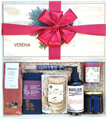 Unsere Winterzauber Geschenkbox beinhaltet köstliche weihnachtliche Zuckermischung, Bio Früchtetee, Schokoladentafel, Sweet Orange Handseife, weihnachtliche Duftkerze, weihnachtliche Seife und eine Schokoladentafel. Auf dem Bild sieht du oben eine geschlossene Box mit Namens-Gravur und grüner Schleife verpackt. Darunter siehst du die gleiche Boxen offen ohne Decke mit den genannten Produkten in die Box gelegt.
