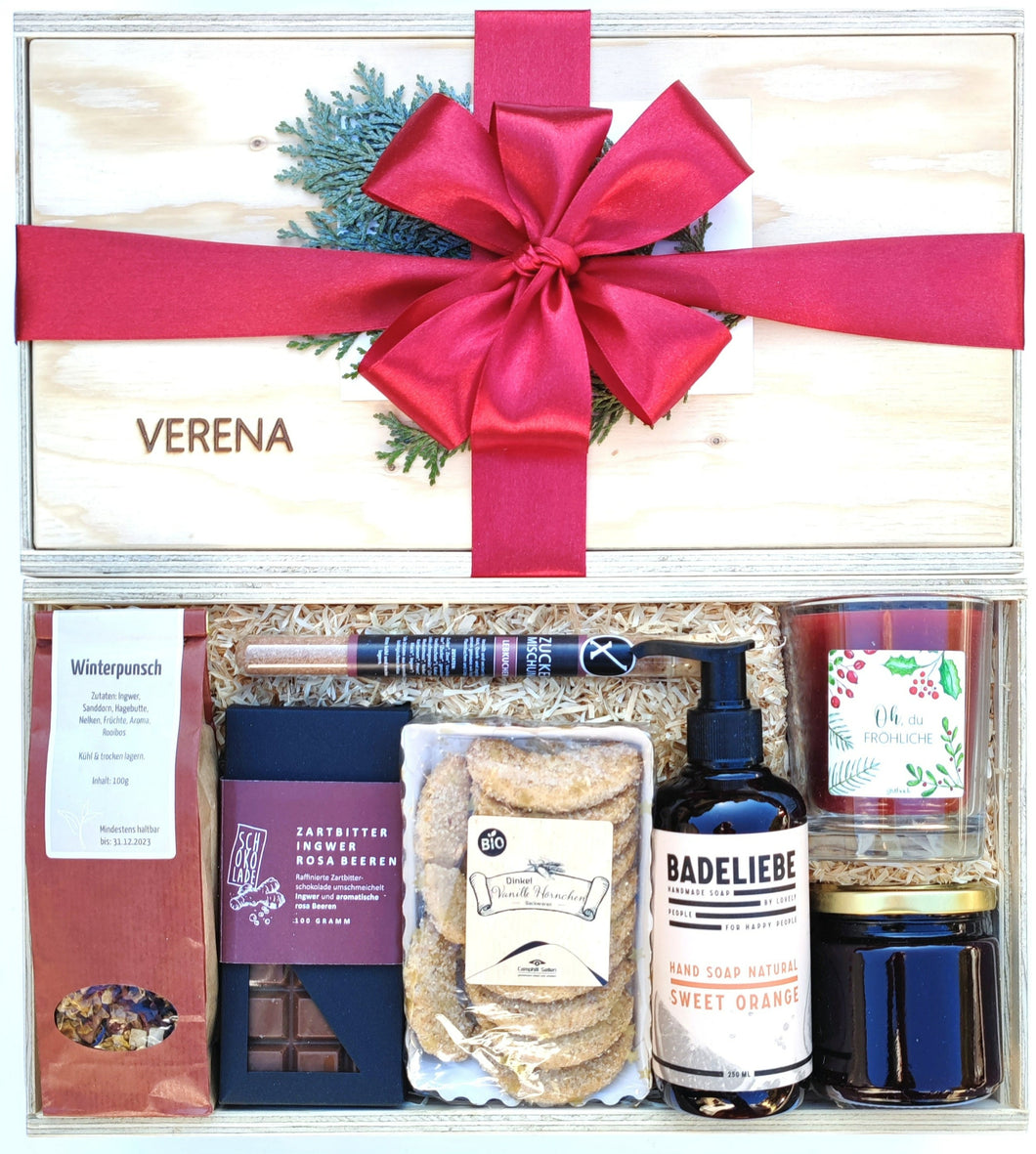 Unsere Winterzauber Geschenkbox beinhaltet köstliche weihnachtliche Zuckermischung, Bio Früchtetee, Schokoladentafel, Sweet Orange Handseife, weihnachtliche Duftkerze, weihnachtliche Seife und eine Schokoladentafel. Auf dem Bild sieht du oben eine geschlossene Box mit Namens-Gravur und grüner Schleife verpackt. Darunter siehst du die gleiche Boxen offen ohne Decke mit den genannten Produkten in die Box gelegt.