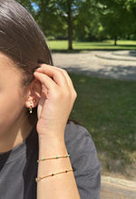 Lade das Bild in den Galerie-Viewer, Auf dem Bild ist eine Frau zu sehen, die ihre Haare hinter´s Ohr steckt. Dabei präsentiert sie ihre Ohrringe und ihre Armbänder. Sie trägt einen goldenen Hängeohrring und einen goldenen Ohrstecker. An ihrem Hängeohrring hängt eine kleine goldene Kugel. An ihrem Arm trägt sie zwei goldene Armketten. An der ersten Armkette sind türkise Steinchen und an dem zweiten lila Steinchen zu finden.
