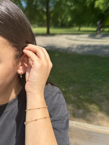 Auf dem Bild ist eine Frau zu sehen, die einen goldenen Ohrstecker und einen goldenen Hänge-Ohrring trägt. An dem Hänge-Ohrring hängt eine goldene kleine Kugel. Die Frau trägt außerdem zwei Armbänder. Die Armbänder bestehen aus goldenen Armketten mit Steinchen. Das erste Armband besitzt türkise Steinchen, das zweite besitzt lila Steinchen.