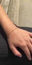 Laden und Abspielen von Videos im Galerie-Viewer, Die Frau sitzt und kreuzt ihre Beine. Ihr Arm liegt auf ihrem Bein.An ihrem Arm trägt sie eine goldene Armkette mit lila Steinchen.
