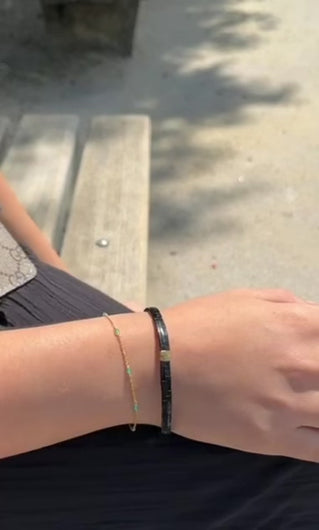 Im Video ist eine Hand mit Armbändern zu sehen. Die Frau trägt zwei verschiedene Armbänder. Das erste Armband ist schwarz und dick. Sie besteht nur aus schwarzen, rechteckigen, flachen Steinchen. In der Mitte stechen zwei goldene Steinchen auf. Das zweite Armband besteht aus einer goldenen Armkette mit türkisen Steinchen.