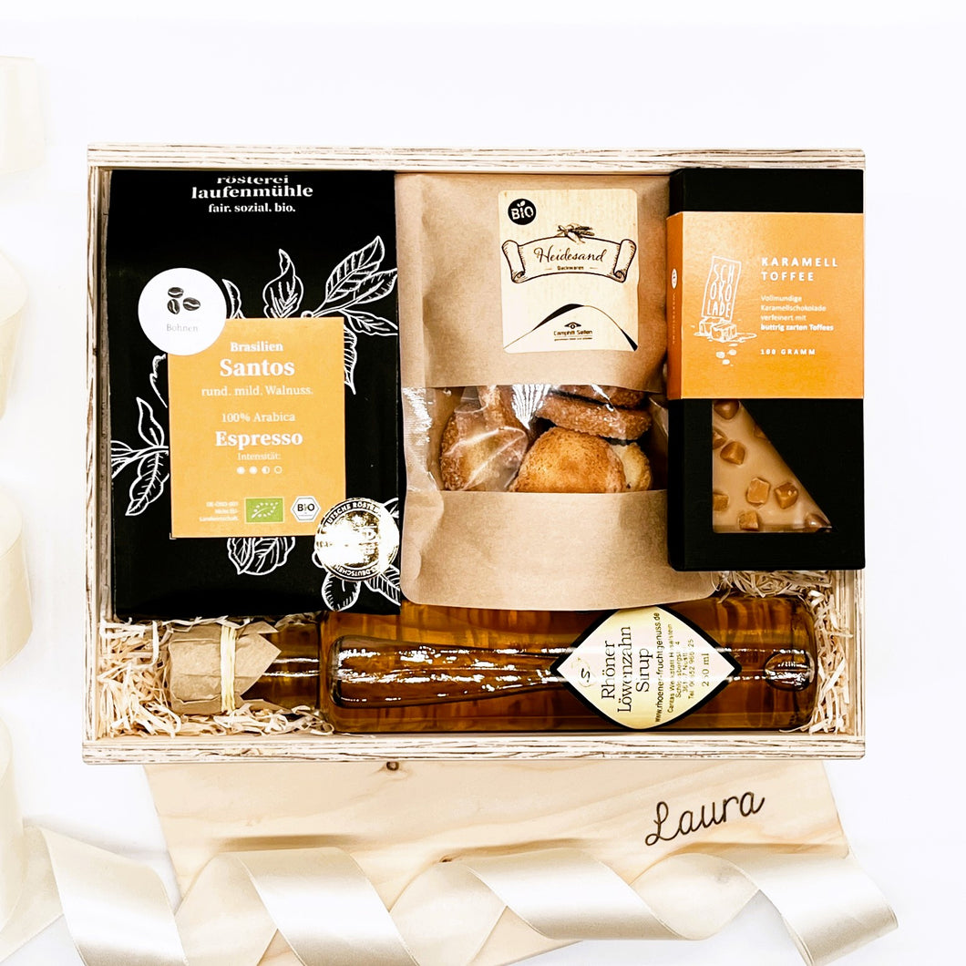 Unsere Geschenkboxen sind immer ein gutes Geschenk für jeden Anlass. Unsere Feinschmecker Box ist hier abgebildet mit Bio Espresso, Heidesand Kekse, Schokolade und Löwenzahnsirup in einer offenen Holzbox.