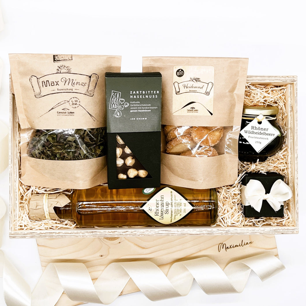 Diese Geschenkbox enthält: Direkt gehandelter Kaffee oder Bio Kräuter Tee (Demeter zertifiziert), Schokoladentafel, Bio Heidesand Kekse, Fruchtaufstrich, Löwenzahn-Sirup und Bio-Schokolade. Hier siehst du ein Bild von der Geschenkbox mit den Produkten in der Box.